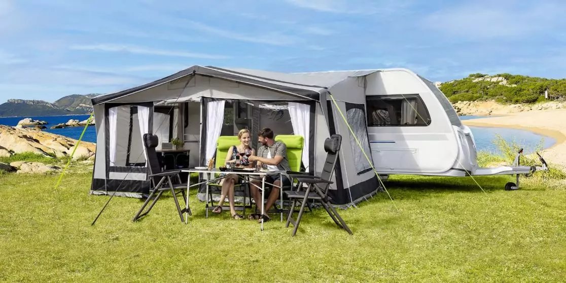 Wohnwagen mit angebautem Vorzelt auf Gras nahe Strand; zwei Personen sitzen an einem Tisch unter dem Vorzelt, au&szlig;en St&uuml;hle und Tisch, Sand, Meer und blauer Himmel im Hintergrund
