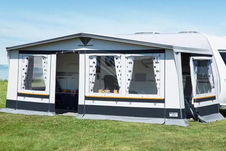 Vorzelt an wei&szlig;em Wohnwagen auf gr&uuml;ner Wiese, drei gro&szlig;e Fenster mit Vorh&auml;ngen und offene Eingangs&ouml;ffnung; Innenraum teilweise sichtbar