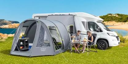 Wei&szlig;es Wohnmobil auf gr&uuml;nem Gras am Meer; graues Vorzelt angebracht; zwei Personen sitzen an einem Campingtisch mit St&uuml;hlen; Regal mit Gegenst&auml;nden im Vorzelt sichtbar; blauer Himmel und felsige K&uuml;ste im Hintergrund