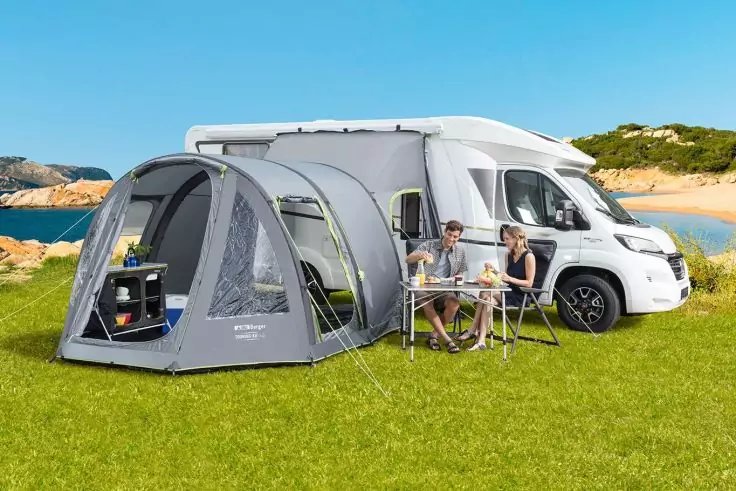 Wei&szlig;es Wohnmobil auf gr&uuml;nem Gras am Meer; graues Vorzelt angebracht; zwei Personen sitzen an einem Campingtisch mit St&uuml;hlen; Regal mit Gegenst&auml;nden im Vorzelt sichtbar; blauer Himmel und felsige K&uuml;ste im Hintergrund
