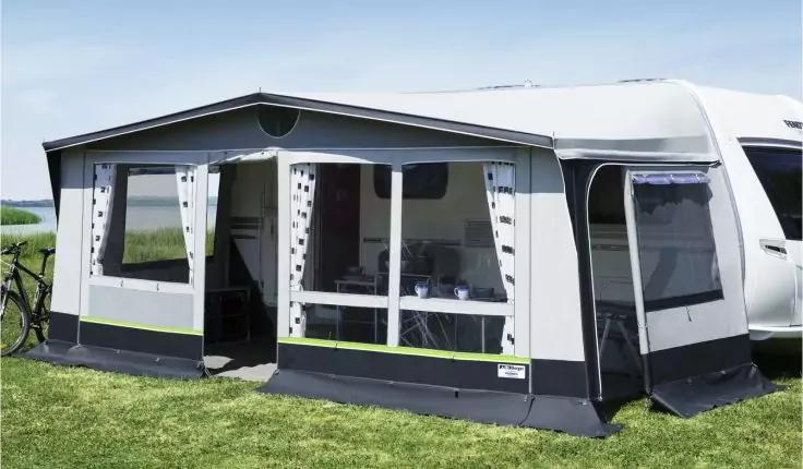 Graues Vorzelt an einem wei&szlig;en Wohnwagen auf Wiese, mit gro&szlig;en transparenten Fenstern und links ein Fahrrad sichtbar