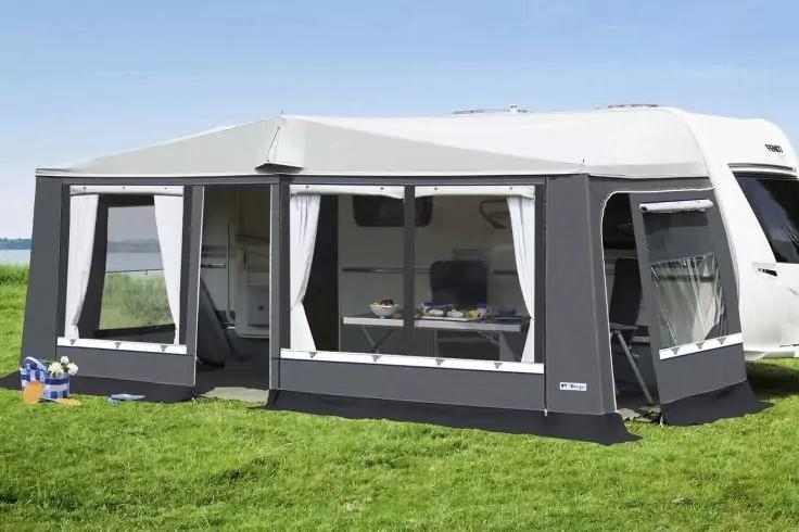 Wei&szlig;er Wohnwagen mit angebautem dunkelgrauen Vorzelt mit Fenstern und wei&szlig;en Vorh&auml;ngen, Tisch und St&uuml;hle sichtbar; gr&uuml;ner Rasen mit kleinen Spielzeugen links, Wasser im Hintergrund und blauer Himmel