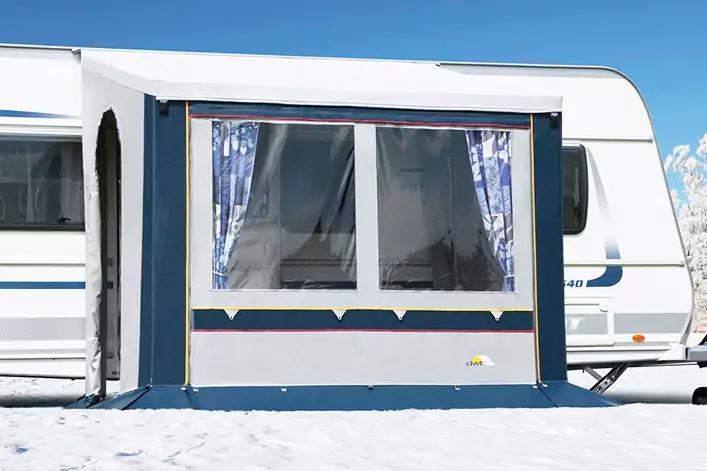 Wei&szlig;er Wohnwagen mit blau-wei&szlig;em angebautem Vorzelt, gro&szlig;es Fenster mit gemusterten Vorh&auml;ngen, verschneiter Boden und blauer Himmel