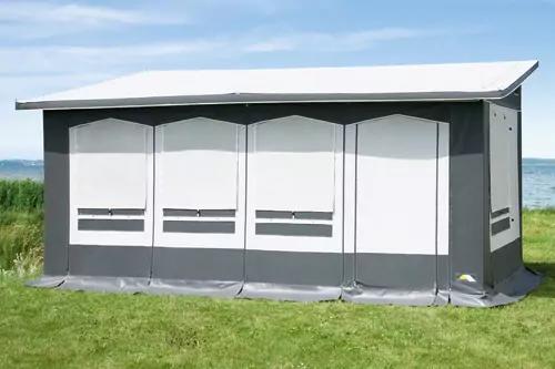 Graue, rechteckige Au&szlig;enstruktur mit drei wei&szlig;en vertikalen Paneelen und flachem Dach auf gr&uuml;ner Wiese, dahinter Wasser und blauer Himmel