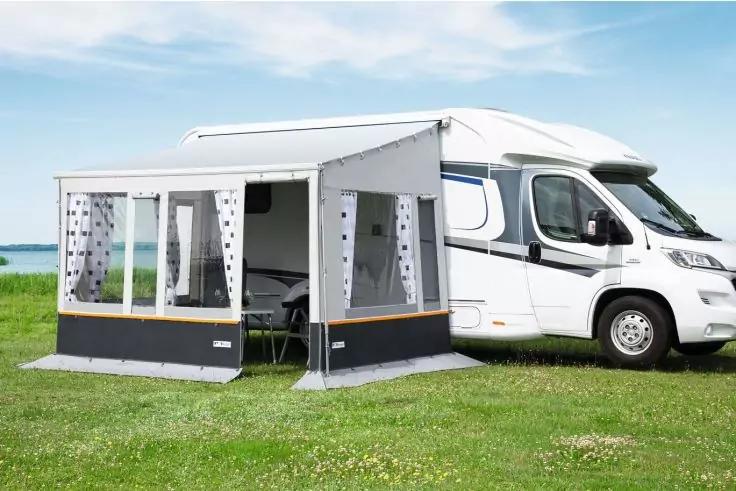 Wei&szlig;es Wohnmobil auf einer Wiese mit angebautem Vorzelt mit Fenstern; im Hintergrund Wasser und blauer Himmel