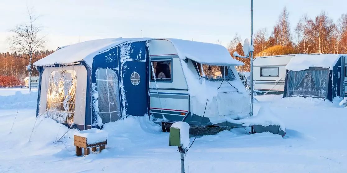 Wei&szlig;er Wohnwagen mit blauem Vorzelt auf verschneitem Boden, weiterer Wohnwagen im Hintergrund, B&auml;ume und Himmel