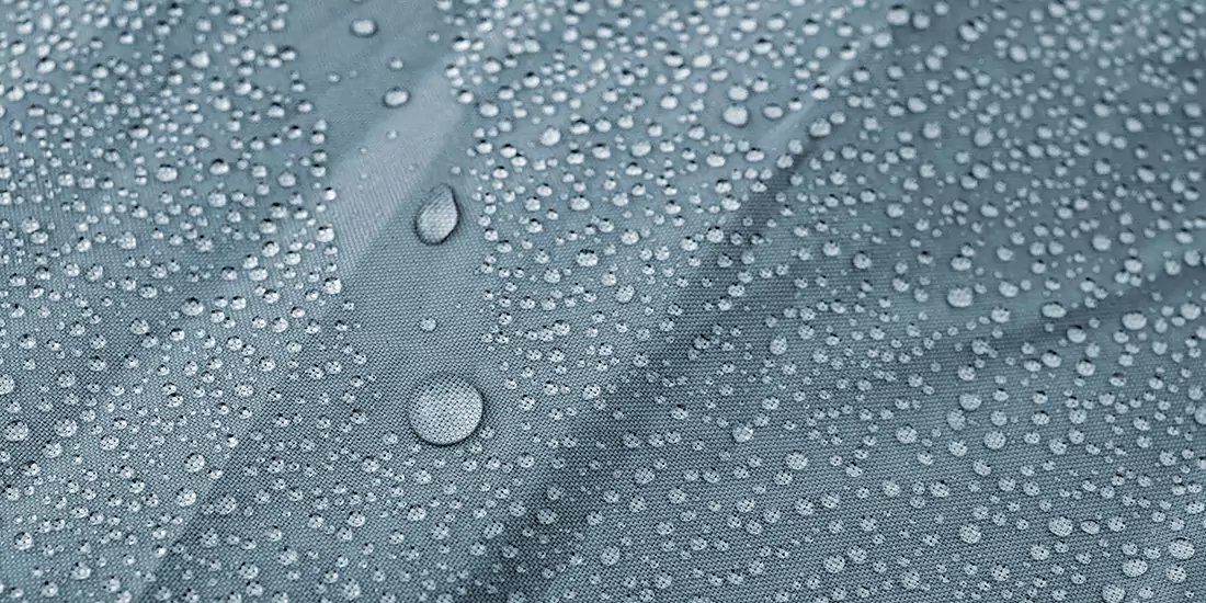 Nahaufnahme von Wassertropfen unterschiedlicher Gr&ouml;&szlig;e auf einer blaugrauen, leicht strukturierten Oberfl&auml;che