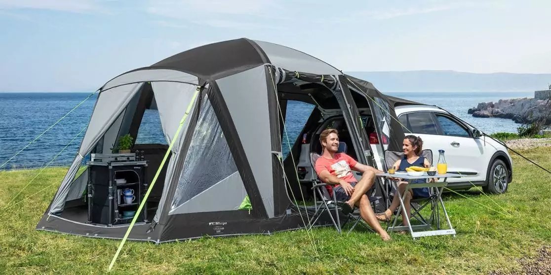 Zwei Personen sitzen auf Campingst&uuml;hlen unter einem grauen Vorzelt, das an ein wei&szlig;es Auto grenzt, mit einem Klapptisch mit Getr&auml;nken und Sch&uuml;ssel; Gras im Vordergrund und K&uuml;ste im Hintergrund