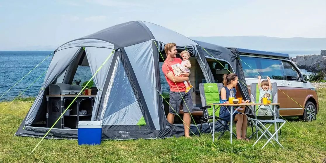 Graues Vorzelt an braun-beigeem Wohnmobil auf einer Wiese am Meer, davor Campingtisch, Campingst&uuml;hle und K&uuml;hlbox. Drei Personen: eine Person h&auml;lt ein Kleinkind, eine sitzt am Tisch, ein Kind steht neben dem Tisch