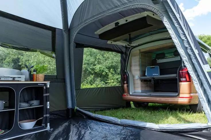 Ge&ouml;ffneter Heckbereich eines orangefarbenen Campers unter grauem Vorzelt: Regale mit Geschirr und einer blauen Box; Wiese und B&auml;ume im Hintergrund