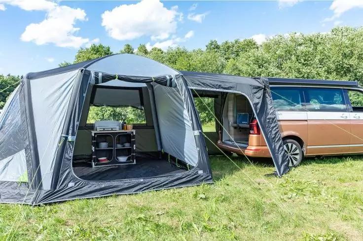 Braun-blauer Camper-Van mit angebautem grauem Vorzelt, ge&ouml;ffnetes Innenzelt mit Regalen und Beh&auml;ltern, Wiese, B&auml;ume und wolkiger Himmel