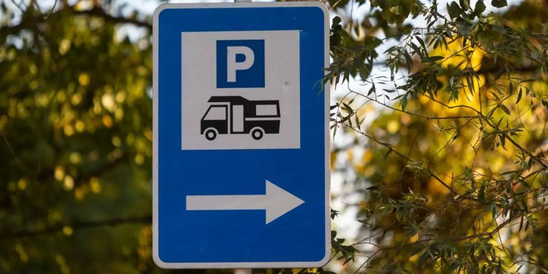 Blaues Schild mit wei&szlig;em &sbquo;P&lsquo;, Wohnmobil-Symbol und wei&szlig;em Pfeil nach rechts, vor B&auml;umen