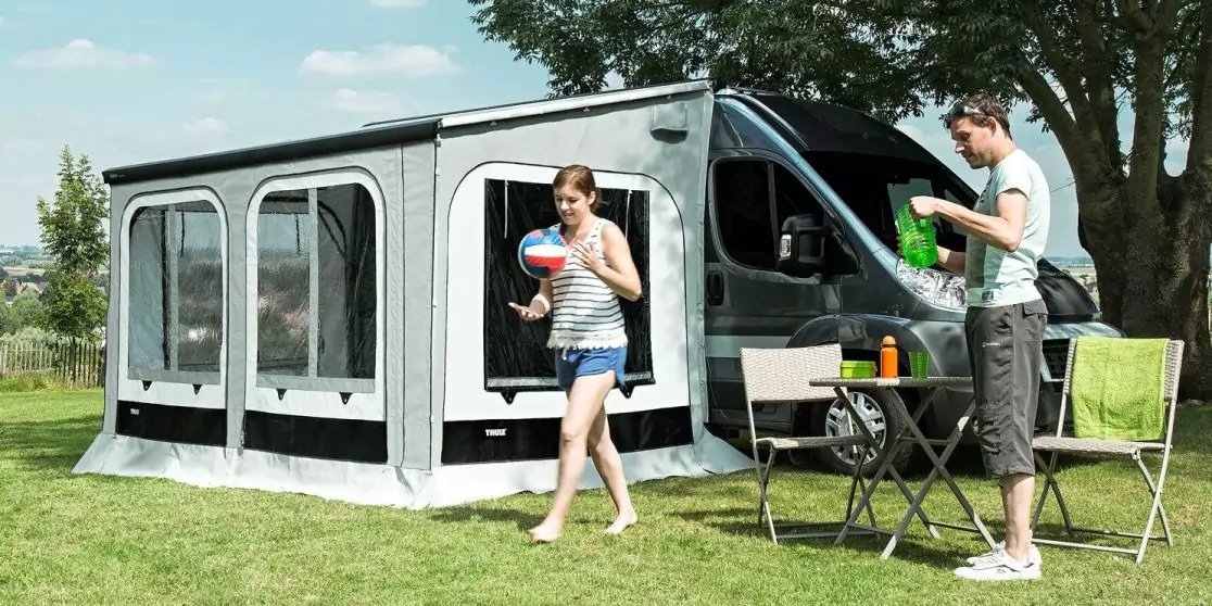 Grauer Van mit angebautem grauem Vorzelt auf Gras; zwei Personen, eine in gestreiftem Top tr&auml;gt einen bunten Ball, die andere steht an einem Klapptisch und h&auml;lt eine gr&uuml;ne Packung; Tisch mit Flaschen und St&uuml;hlen; Baum und Himmel im Hintergrund