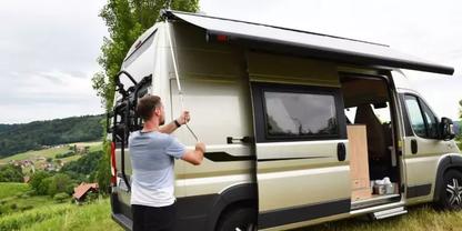 Wohnmobil auf Wiese, seitliche T&uuml;r offen, Person klappt Markise aus, Fahrrad am Heck, Dorf und H&uuml;gel im Hintergrund