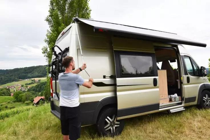 Wohnmobil auf Wiese, seitliche T&uuml;r offen, Person klappt Markise aus, Fahrrad am Heck, Dorf und H&uuml;gel im Hintergrund