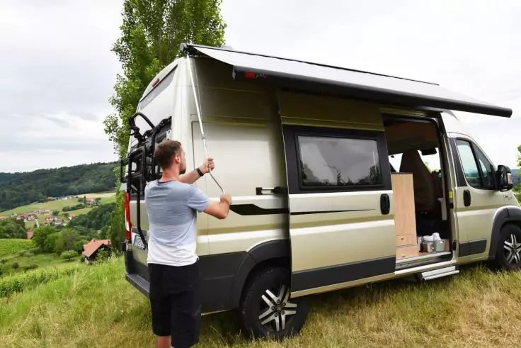 Wohnmobil auf Wiese, seitliche T&uuml;r offen, Person klappt Markise aus, Fahrrad am Heck, Dorf und H&uuml;gel im Hintergrund