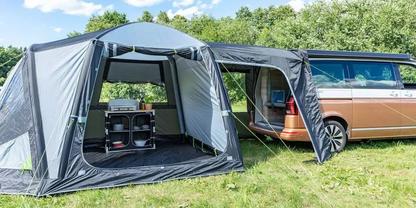 Braun-wei&szlig;er Campingbus mit angebautem grauem Vorzelt auf einer Wiese, offenes Vorzelt mit Regalen, B&auml;ume im Hintergrund