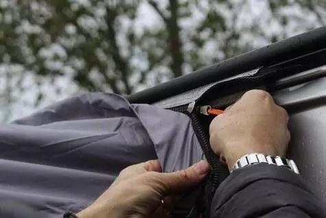 H&auml;nde an einem grauen Stoff mit Rei&szlig;verschluss; orangefarbener Rei&szlig;verschlusszug, silberne Armbanduhr und verschwommene B&auml;ume im Hintergrund