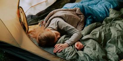 Person liegt auf der Seite in einem ge&ouml;ffneten Zelt auf einem Schlafsack, tr&auml;gt graues Oberteil, blondes Haar