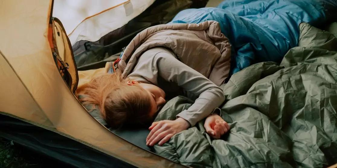 Persoon ligt op de zij in een open tent op een slaapzak, draagt een grijs bovenstuk, blond haar