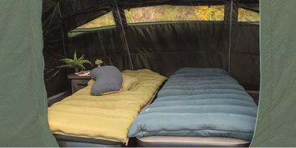 Zwei nebeneinander liegende Schlafmatratzen mit Decken (links gelb, rechts blau) und Kissen in einem aufgebauten Zelt; links ein kleiner Beistelltisch mit einer Pflanze