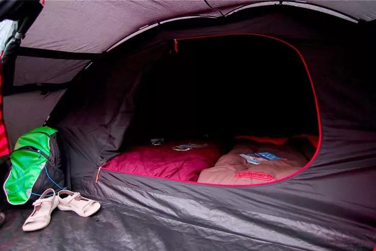 Innenansicht eines Zelts mit offenem Eingang, zwei Schlafs&auml;cken (rot und beige) auf dem Boden, links helle Sandalen und eine gr&uuml;ne Tasche