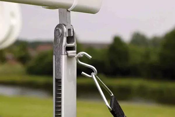 Nahaufnahme eines Metallkarabiners, befestigt an einer Metallhalterung mit schwarzem Gurt; unscharfer Hintergrund mit Wiese, B&auml;umen und Wasser