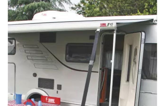 Wei&szlig;er Wohnwagen mit ausgefahrener Markise, ge&ouml;ffneter T&uuml;r, seitlichem Fenster, L&uuml;ftungsgitter und roter Kunststoffkiste im Vordergrund; B&auml;ume im Hintergrund