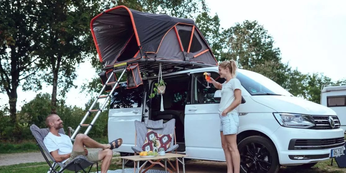 Wei&szlig;er Campervan mit aufgeklapptem Dachzelt und Leiter, offene Seitent&uuml;r, Mann sitzt in Campingstuhl, Frau steht neben dem Fahrzeug, kleiner Campingtisch mit Geschirr, B&auml;ume und Rasen im Hintergrund