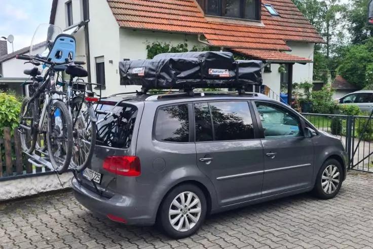 Graues Auto mit schwarzer Dachtasche und Fahrradtr&auml;ger am Heck mit zwei Fahrr&auml;dern, geparkt vor einem wei&szlig;en Haus mit rotem Ziegeldach