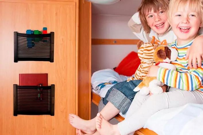 Zwei l&auml;chelnde Kinder sitzen auf einem Bett, umarmen sich und halten Kuscheltiere; neben dem Bett ein Holzpaneel mit Aufbewahrungstaschen