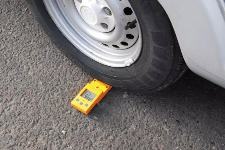 Weißes Fahrzeugrad auf grauem Asphalt, gelbes elektronisches Gerät mit Display liegt teilweise unter dem Reifen