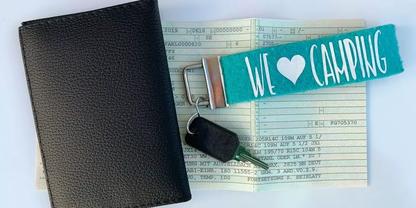 Schwarze H&uuml;lle, gefaltete Papiere mit gedrucktem Text, Autoschl&uuml;ssel mit schwarzem Kunststoffschl&uuml;sselkopf und t&uuml;rkisfarbenem Schl&uuml;sselband mit wei&szlig;er Aufschrift WE &hearts; CAMPING