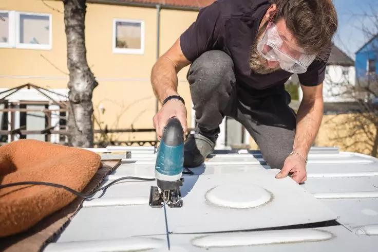 Person kniet auf Metall&shy;dach und s&auml;gt mit einer elektrischen Stichs&auml;ge entlang einer markierten Linie; tr&auml;gt Schutzbrille; braunes Polster, runde L&uuml;ftungs&ouml;ffnung und H&auml;user im Hintergrund