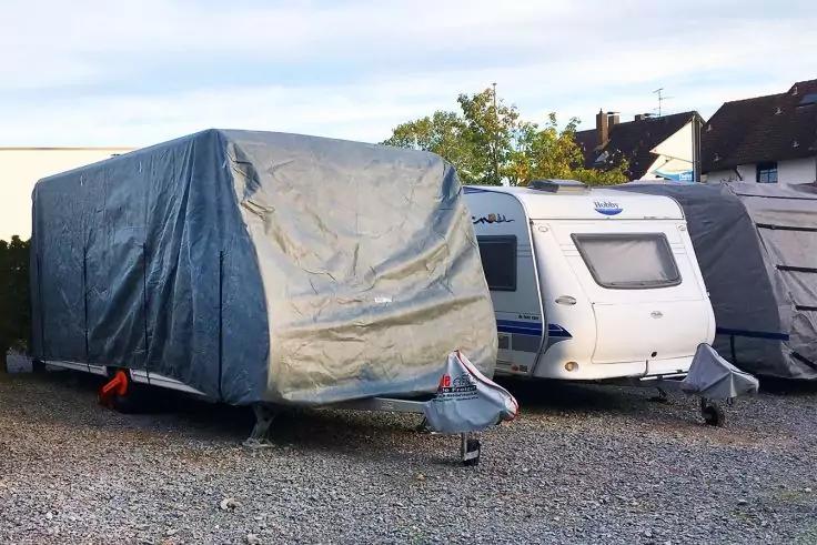 Zwei Wohnwagen auf einem Schotterplatz, einer mit grauer Plane bedeckt, der andere weiß, Häuser im Hintergrund
