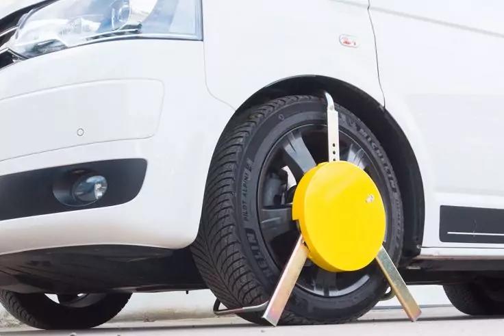 Voorzijde links van een wit voertuig, voorwiel links met een gele wielklem en drie metalen armen