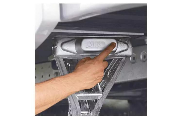 Hand mit ausgestrecktem Zeigefinger drückt einen rechteckigen Knopf an einer metallischen Scharnier-/Befestigungsmechanik unter einer Decke