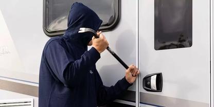 Person mit dunkler Kapuzenjacke h&auml;lt ein Metallwerkzeug an die T&uuml;r eines Wohnmobils; zwei Fenster sichtbar