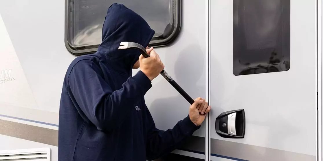 Person mit dunkler Kapuzenjacke h&auml;lt ein Metallwerkzeug an die T&uuml;r eines Wohnmobils; zwei Fenster sichtbar