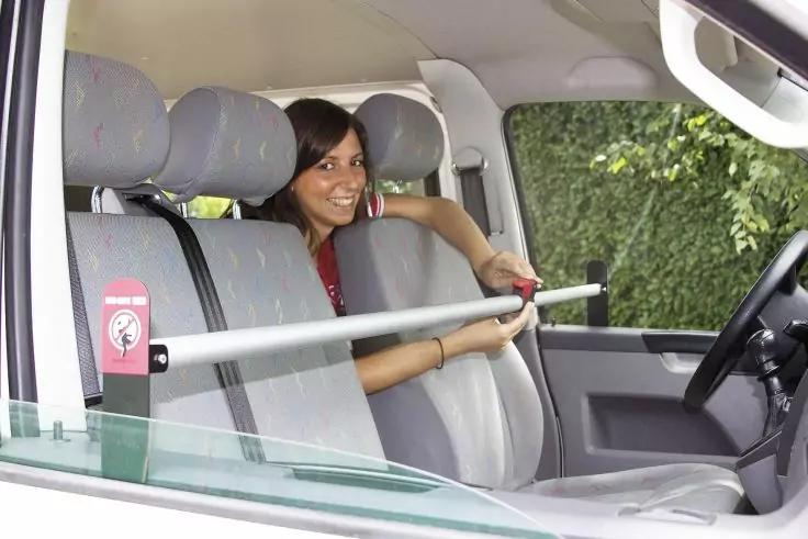 Frau sitzt im Fahrzeug hinter den Vordersitzen und hält eine horizontale Metallstange; Lenkrad und grünes Laub am Fenster sichtbar
