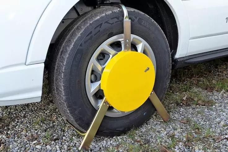 Gelbes rundes Radschloss mit drei Metallstreben an einem Reifen eines weißen Fahrzeugs auf Schotter