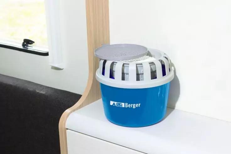 Blauer Plastikbeh&auml;lter mit wei&szlig;em Gitterrand und grauem Deckel, Aufdruck 'Berger', steht auf einer wei&szlig;en Ablage neben hellen Holzleisten und einem Fenster; links ein schwarzes Sitzkissen