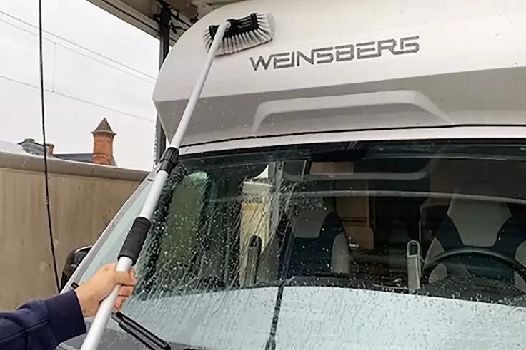 Hand h&auml;lt eine Teleskopstange mit B&uuml;rste an einer nassen Windschutzscheibe eines Wohnmobils mit Schriftzug WEINSBERG, im Hintergrund Hauswand und Dach sichtbar