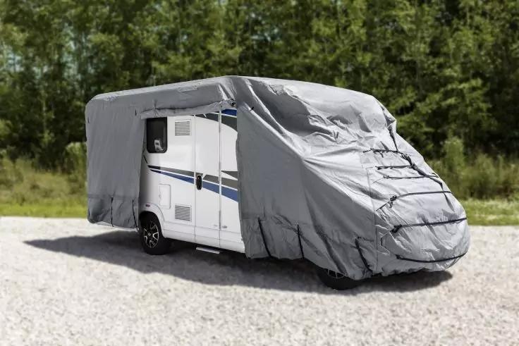 Wei&szlig;es Wohnmobil mit blauen Streifen, gr&ouml;&szlig;tenteils mit grauer Schutzplane bedeckt, auf Kies vor B&auml;umen