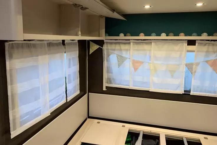 Zimmerecke mit drei Fenstern, weiße transparente Vorhänge, pastellfarbene Wimpelkette am mittleren Fenster, türkisfarbene Ablage mit runden Dekorationsgegenständen darüber, Einbauleuchten in der Decke, sichtbares Lattenrost/Bettgestell unten