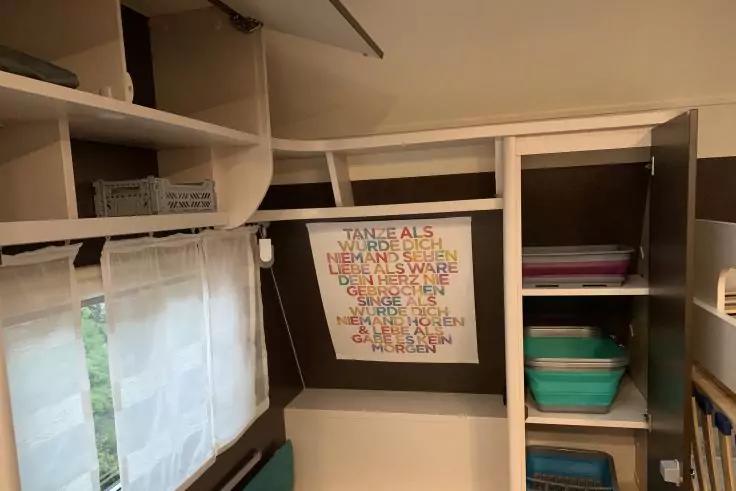 Innenraum mit weißen Regalen und Schränken; links ein Fenster mit weißem halbtransparentem Vorhang, darunter ein türkisfarbenes Sitzkissen; an der Rückwand ein gerahmtes buntes Textbild; rechts Regale mit gestapelten Kunststoffboxen (türkis, rosa)