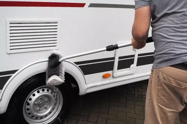 Teilweise sichtbare Person von hinten in grauem T‑Shirt und beigen Shorts, hält eine lange Bürste an der Seitenwand eines weißen Wohnwagens über dem Rad; sichtbarer Reifen, orange Positionsleuchte, gepflasterter Untergrund