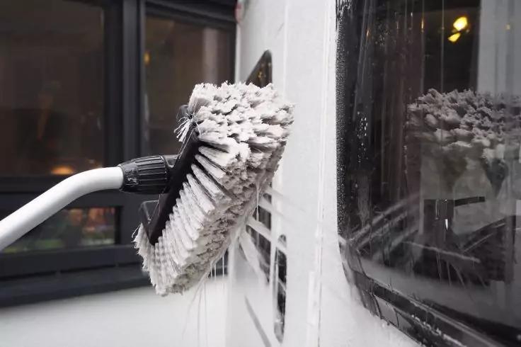 Reinigungsbürste mit Schaum an einem weißen Fensterrahmen, Seifenwasser läuft am Glas herunter