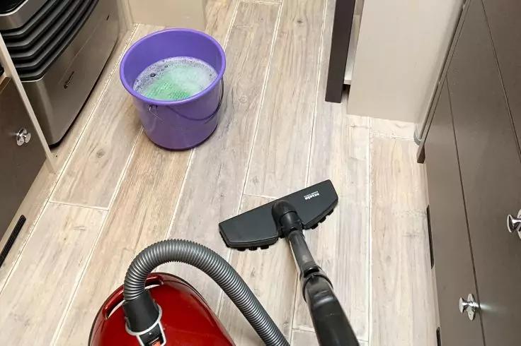 Roter Bodenstaubsauger mit Schlauch und Bodendüse auf hellem Holzoptikboden; lila Eimer mit schäumendem Wasser oben links; graue Schränke rechts