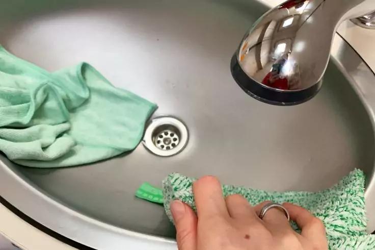 Hand mit Ring hält grünes Reinigungstuch in einer Edelstahlspüle; Chromarmatur und Abfluss sichtbar